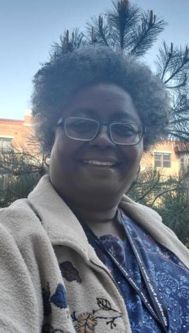 Photo of Kim, a smiling black woman with curly gray and black hard wearing a blue shirt and a beige jacket embroidered with vines and leaves, stands in front of a small pine tree.